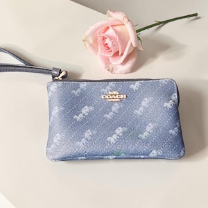 May include: A blue Coach wristlet featuring a pattern of white horse-drawn carriages. It has a zip closure, a small gold Coach logo, and a blue wrist strap. A pink rose is placed nearby.