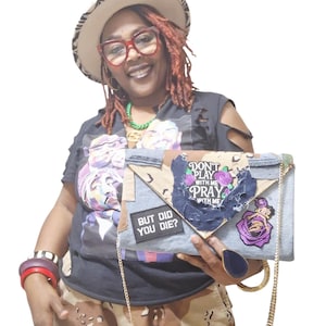 Puede incluir: Un bolso de mano de mezclilla con una correa de cadena dorada, con parches con texto y diseños florales. El bolso de mano tiene un cierre de solapa y lo sostiene una persona que lleva un sombrero y gafas.