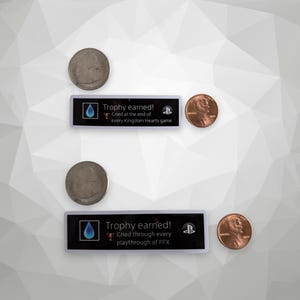 以下が含まれることがあります： 「Trophy earned!」の文字と青い涙のアイコンが入った、長方形の白黒ゲームトロフィーが2つ。トロフィーには「Cried at the end of every Kingdom Hearts game」と「Cried through every playthrough of FFX」と書かれています。2つの25セント硬貨と2つの1セント硬貨も画像にあります。