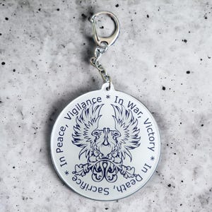 May include: A silver-coloured round keyring with a chain and clasp. The keyring features a detailed emblem of a bird with outstretched wings, surrounded by the words "In Peace, Vigilance, In War, Victory, In Death, Sacrifice."