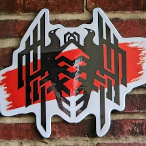 May include: A white, die-cut sticker with a black and red graphic design. The design features two stylized birds above a geometric pattern, with a red brushstroke across the center. The sticker is mounted on a brick wall.
