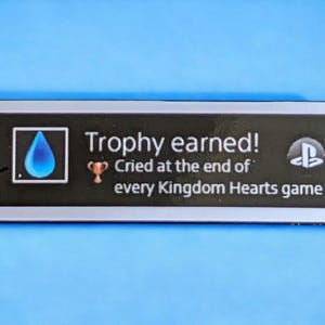以下が含まれることがあります： 「Trophy earned! Cried at the end of every Kingdom Hearts game」のテキストが入った、白い縁取りのある黒い長方形のステッカー。青い涙のアイコン、トロフィーアイコン、PlayStationのロゴが特徴です。
