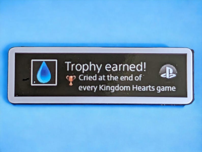以下が含まれることがあります： 「Trophy earned! Cried at the end of every Kingdom Hearts game」のテキストが入った、白い縁取りのある黒い長方形のステッカー。青い涙のアイコン、トロフィーアイコン、PlayStationのロゴが特徴です。