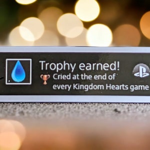 以下が含まれることがあります： 「Trophy earned! Cried at the end of every Kingdom Hearts game」のテキストが入った、長方形の白黒ピン。青い涙のアイコンとPlayStationのロゴも特徴です。