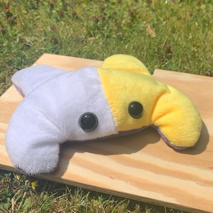 May include: A plush toy shaped like a creature with a white and yellow body, black eyes, and a purple mouth. The toy is lying on a wooden surface.