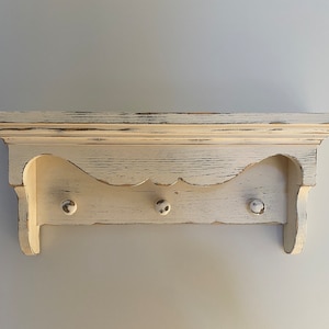 May include: White wooden shelf with a scalloped edge and three knobs. The shelf has a distressed finish.