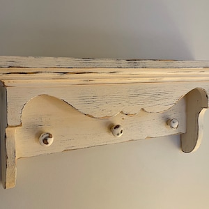 May include: White wooden shelf with a scalloped edge and three knobs. The shelf has a distressed finish, giving it a vintage look.
