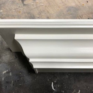 May include: A section of white painted crown molding. The molding has a layered design with a curved edge. The piece is angled, showing the detailed profile and the smooth, painted surface. The molding is likely for interior design.