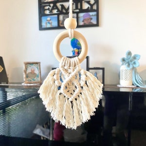 May include: A white macrame hanging with a wooden ring and a wooden bead. The macrame has a fringe and is in the shape of a teardrop.