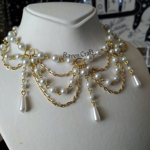 May include: A statement necklace featuring faux pearls and gold-tone chain. The necklace has multiple strands of pearls and chains, with teardrop-shaped pearl pendants. The Raven.Craft logo is visible on the display.