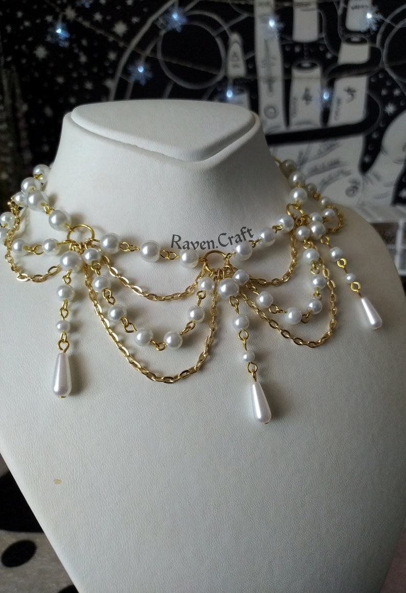 May include: A statement necklace featuring faux pearls and gold-tone chain. The necklace has multiple strands of pearls and chains, with teardrop-shaped pearl pendants. The Raven.Craft logo is visible on the display.