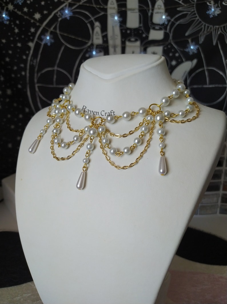 May include: A gold-toned necklace with multiple strands of faux pearls and delicate chains. The necklace features a cascading design with teardrop-shaped pearl pendants. The Raven.Craft logo is visible on the necklace.