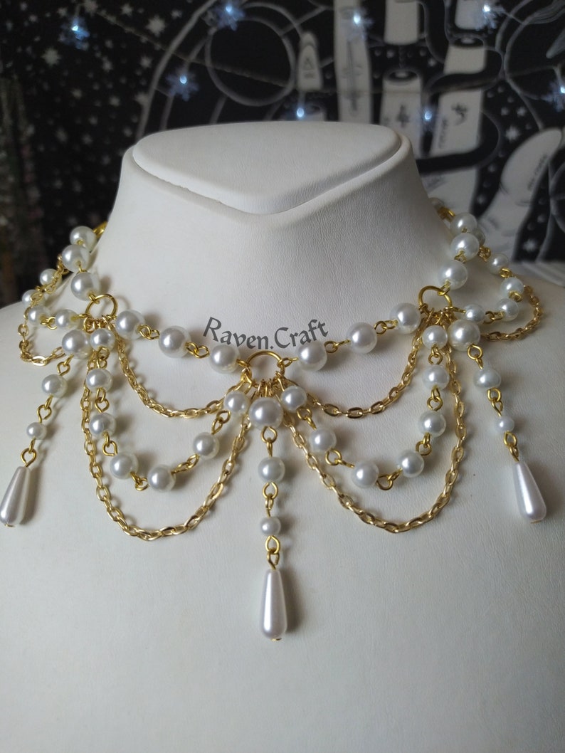 May include: A pearl and gold-tone necklace with the text "Raven Craft". The necklace features multiple strands of faux pearls and gold-colored chain, with teardrop-shaped pearl pendants.