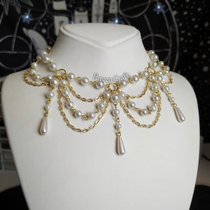May include: A pearl and gold-tone necklace displayed on a white mannequin. The necklace features multiple strands of small, round pearls and gold-colored chains. The design includes dangling pearl drops. The text "Raven Craft" is visible.