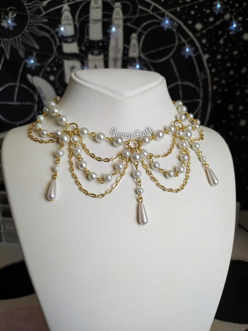 May include: A pearl and gold-tone necklace displayed on a white mannequin. The necklace features multiple strands of small, round pearls and gold-colored chains. The design includes dangling pearl drops. The text "Raven Craft" is visible.