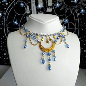 Peut inclure: Un collier doré avec un pendentif en forme de croissant de lune et des perles de cristal bleues pendantes. Le collier comporte de petites étoiles dorées et des perles bleues le long de la chaîne. Le logo Raven Craft est visible.