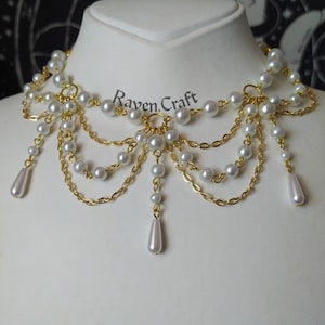 May include: A white pearl and gold chain necklace with a layered design. The necklace features multiple strands of small, round pearls and gold chains, with teardrop-shaped pearl pendants. The text "Raven Craft" is visible.