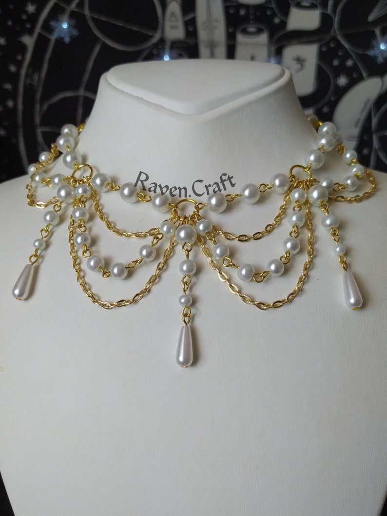May include: A white pearl and gold chain necklace with a layered design. The necklace features multiple strands of small, round pearls and gold chains, with teardrop-shaped pearl pendants. The text "Raven Craft" is visible.