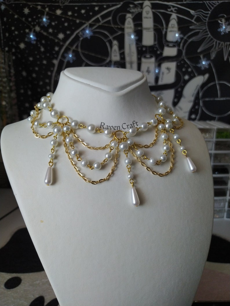 May include: A pearl and gold-tone necklace displayed on a white mannequin. The necklace features multiple strands of small, round white pearls connected by delicate gold chains. Teardrop-shaped pearl pendants hang from the bottom. The text "Raven.Craft" is visible.