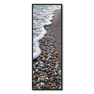 Puede incluir: Impresión vertical enmarcada que representa una playa. La imagen muestra guijarros coloridos y lisos a lo largo de la orilla, con olas blancas y espumosas que los cubren. Los guijarros son de varios colores, incluyendo marrón, azul y naranja.