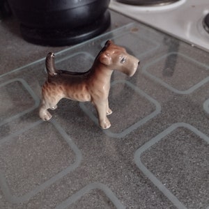 May include: A small, brown and white ceramic figurine of a terrier dog. The figurine is standing on a grey tiled surface.