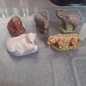 May include: Five ceramic animal figurines, including a brown bear, a grey rhinoceros, a grey elephant, a white polar bear, and a brown and orange tiger. The figurines are all sitting on a grey tiled surface.