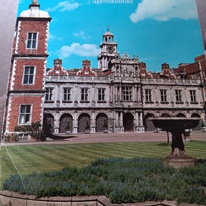 May include: A postcard depicting Hatfield House in Hertfordshire. The image shows the stately home with a clock tower, red brickwork, and formal gardens under a blue sky. The text "HATFIELD HOUSE HERTFORDSHIRE" is displayed at the top of the card.