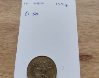 May include: A South Korea 10 Won coin from 1994 featuring a pagoda. The coin is gold in colour and is on a white piece of paper with handwritten text that says "South Korea 10 Won 1994 £1.50".