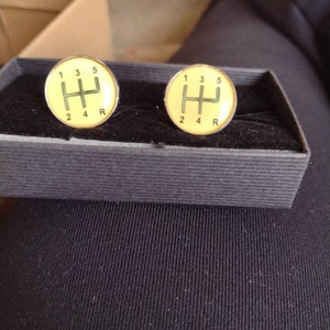 May include: A pair of cufflinks with a yellow background and a silver gear shift pattern. The numbers 1, 2, 3, 4, 5, and R are printed around the gear shift pattern.