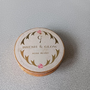 May include: A circular rose blush compact with a white lid, gold detailing, and the text "BRUSH & GLOW" and "ROSE BLUSH". The compact has a gold edge and a pink rose illustration.