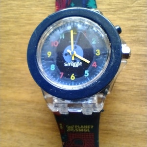 May include: A blue and clear plastic watch with a black face and colourful numbers. The watch band has a colourful graphic print with the text "Planet Smgl" and "Danger".