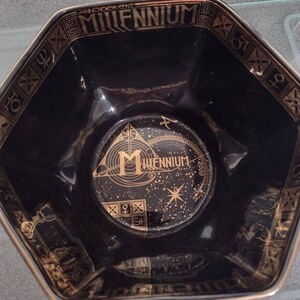 May include: A black octagonal bowl with a gold rim and a gold design featuring the text "Anno Domini Millennium 2000". The bowl has a gold star and a gold triangle with the letter "M" and the text "Millennium 2000" inside.