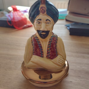 May include: A ceramic figurine of a man wearing a blue turban with a gold jewel on his forehead. He is wearing a red and gold patterned waistcoat and has a black beard. He is sitting with his arms crossed and is on a gold base.