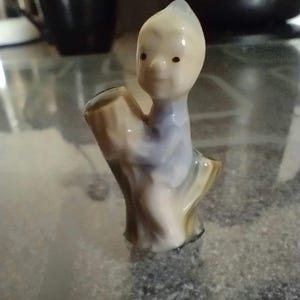 May include: A small porcelain figurine of a boy sitting on a tree stump. The boy is wearing a blue shirt and has a light brown complexion. The tree stump is painted in shades of brown and beige.
