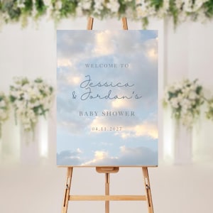 Könnte beinhalten: Ein hölzernes Staffelei-Display zeigt ein Schild mit einem Wolkenhintergrund. Auf dem Schild steht "WELCOME TO Jessica & Jordan's BABY SHOWER 04.11.2027" in eleganter Schrift. Weiße Blumengestecke sind im Hintergrund zu sehen.