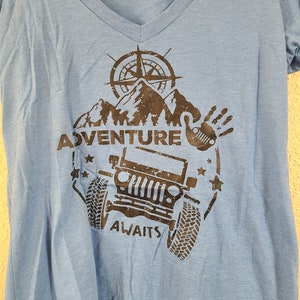 Puede incluir: Una camiseta azul con un diseño gráfico negro que presenta una brújula, montañas, un jeep y el texto "Adventure Awaits".
