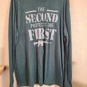 May include: A green long-sleeved t-shirt with a white graphic print that reads "The Second Protects The First" with a stylized image of a rifle underneath the text.