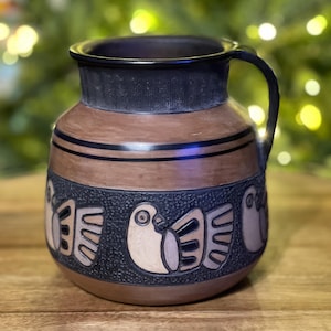 May include: A brown ceramic pitcher with a handle, featuring a black rim and base. The body has a textured design with bird motifs in cream and black bands. The pitcher is on a wooden surface.