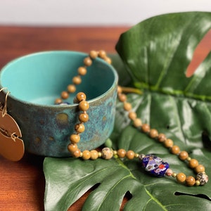 May include: A teal ceramic bowl with a textured finish, a gold-colored earring, and a beaded necklace with a blue and floral-patterned bead. The necklace and bowl are on a green leaf.