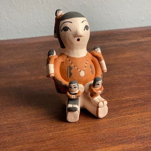 May include: A hand-painted ceramic figurine of a woman with four children. The woman is wearing a brown dress with black and white geometric designs. The children are all wearing brown dresses with black and white geometric designs. The figurine is made of clay and has a rustic, handmade look.