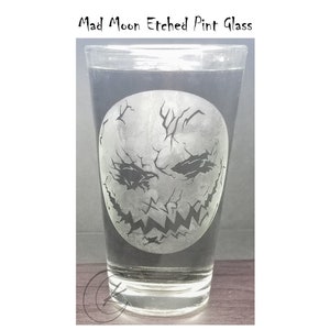 May include: A clear glass pint glass with an etched design of a white, cracked moon face with a sinister grin. The glass is filled with a clear liquid.