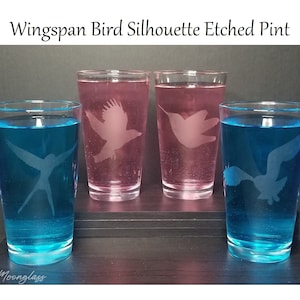 May include: Four etched pint glasses with different bird silhouettes. Two glasses are filled with a pink liquid and two glasses are filled with a blue liquid. The glasses are on a wooden surface.