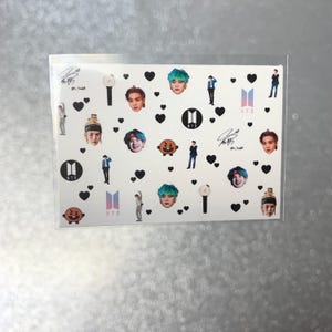 May include: A clear plastic sheet filled with various images of people, hearts, and logos. The images include faces, figures, and the BTS logo. The sheet is on a light gray surface.