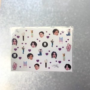 May include: A clear sticker sheet featuring various images of a musical group's members, purple hearts, logos, and signatures. The sticker sheet has a white background.