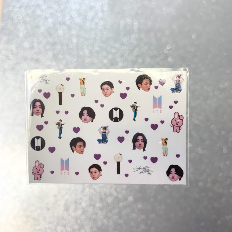 May include: A clear sticker sheet featuring various images of a musical group's members, purple hearts, logos, and signatures. The sticker sheet has a white background.