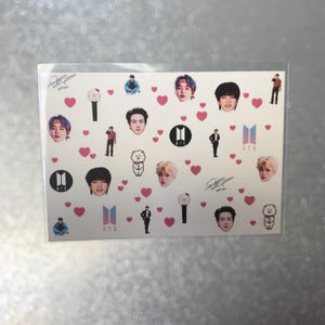 May include: A clear plastic sleeve containing a sheet of stickers. The stickers feature images of people, hearts, logos, and signatures. The stickers are in various colors, including pink, black, white, and blue.