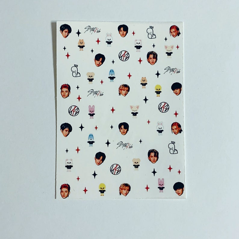 Op de afbeelding: Een vel stickers met de K-pop groep Stray Kids. De stickers zijn wit met zwarte en rode accenten. De stickers bevatten het logo van de groep, foto's van de individuele leden en cartoon dierillustraties.