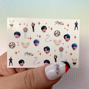 Op de afbeelding: Een vel nagelstickers met afbeeldingen van een K-pop-groep genaamd Stray Kids. De stickers bevatten afbeeldingen van de bandleden, hun logo en een vos-mascotte. De stickers zijn op een witte achtergrond met rode sterren.