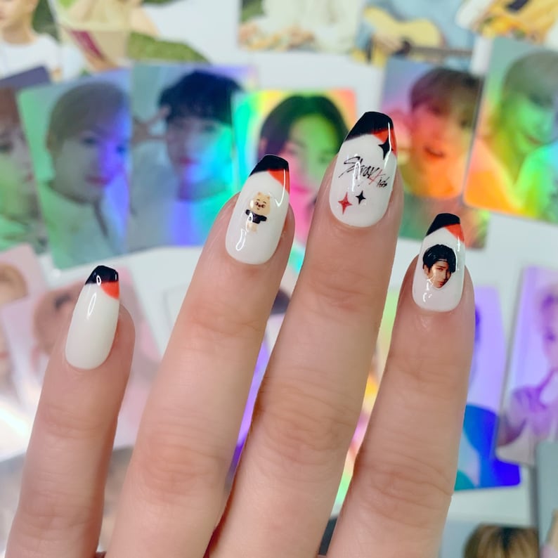 Op de afbeelding: Close-up van een hand met verschillende witte nageltips versierd met zwarte en rode accenten. De nageltips bevatten afbeeldingen van een cartoonbeer, een bandlogo en een gezicht.