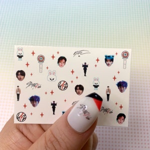 Op de afbeelding: Een vel nagelstickers met een witte achtergrond en verschillende kleurrijke ontwerpen. De ontwerpen omvatten een cartoonkonijn, een microfoon, een ster en een groep mannen. De tekst "Stray Kids" is op het vel gedrukt.
